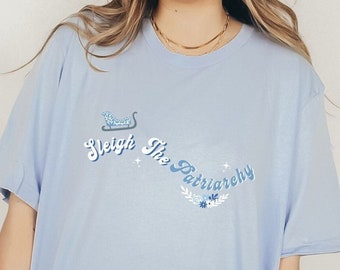 Pode incluir: T-shirt azul claro com a frase "Sleigh The Patriarchy" em uma fonte lúdica. O design inclui um trenó e detalhes florais em tons de azul. Uma peça confortável e casual.