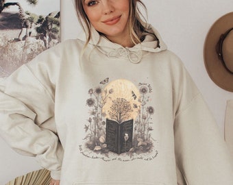 May include: A beige hoodie with a graphic print of a book, a tree, a moon, and butterflies. The text on the graphic reads "Once upon a time... with freedom, laughter, memories, and the moon, she could be happy."