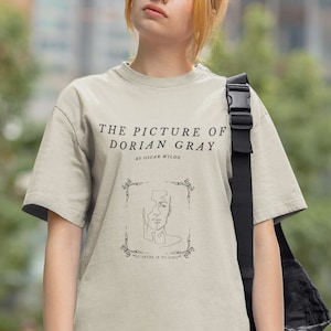 May include: A beige t-shirt with a black graphic print of the book cover for "The Picture of Dorian Gray" by Oscar Wilde. The text "To define is to limit" is printed below the image.