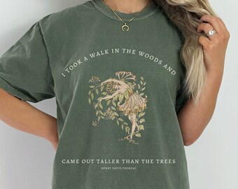 May include: A green t-shirt featuring a vintage-style illustration of a woman in a forest. The text on the shirt reads "I took a walk in the woods and came out taller than the trees - Henry David Thoreau".