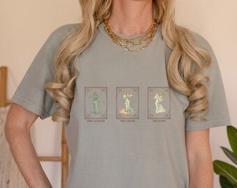 Puede incluir: Una camiseta azul claro con tres ilustraciones de mujeres en marcos de estilo vintage. El texto debajo de cada marco dice "The Lunatic", "The Lover" y "The Poet".