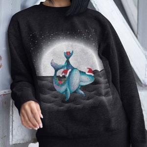 May include: A black sweatshirt with a Christmas-themed design featuring two whales wearing Santa hats in front of a full moon. The whales are surrounded by a starry night sky and a wavy ocean.