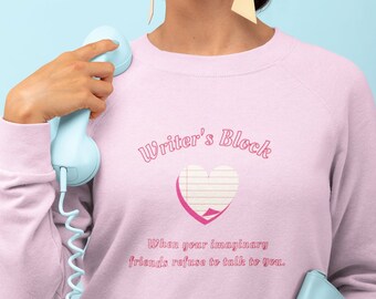 May include: A pink sweatshirt with the text "Writer's Block" and a pink heart with a lined paper design. The text below the heart reads "When your imaginary friends refuse to talk to you."