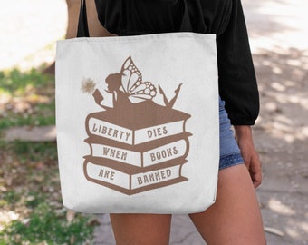 May include: White tote bag with a brown silhouette of a fairy sitting on a stack of books. The books have the text "Liberty Dies When Books Are Banned".