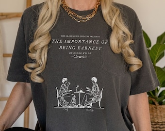 Puede incluir: Una camiseta gris con un estampado gráfico blanco de dos mujeres sentadas en una mesa con el texto "The Importance of Being Earnest by Oscar Wilde" sobre la imagen.