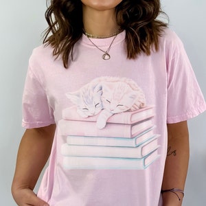 May include: A pink t-shirt with a graphic of two kittens sleeping on a stack of books. The kittens are white and light blue.