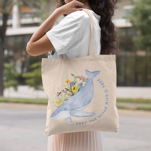 May include: A beige tote bag featuring a blue whale illustration and colourful flowers. The text "Keep the ocean wild & free" is printed on the bag.