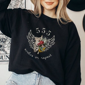 Könnte beinhalten: Schwarzer Pullover mit einem weißen Grafikdruck eines Brustkorbs mit Flügeln und Blumen. Der Text "555 evolve or repeat" ist unter dem Grafikdruck aufgedruckt.