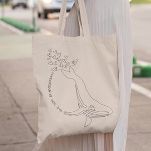 Pode incluir: Uma bolsa tote bege com um desenho de linha preta de uma baleia cercada de corações. O texto "overwhelmed with love" está escrito na bolsa.
