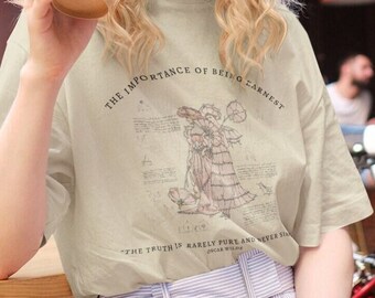 Puede incluir: Una camiseta verde claro con una ilustración vintage de una mujer con un vestido y el texto "THE IMPORTANCE OF BEING EARNEST" y "THE TRUTH IS RARELY PURE AND NEVER SIMPLE" con el nombre "OSCAR WILDE" debajo.