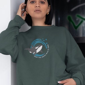 Könnte beinhalten: Ein dunkelgrüner Pullover mit einem Aufdruck einer Orca-Wals, die in einer blauen Welle schwimmt. Der Text "Keep the Orcas Wild & Free" ist um den Aufdruck herum gedruckt.