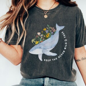 以下が含まれることがあります： 青いクジラとカラフルな花が描かれたダークグレーのTシャツ。Tシャツには「Keep the ocean wild & free」という文字がプリントされています。