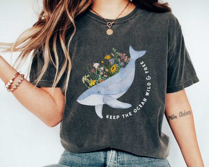 以下が含まれることがあります： 青いクジラとカラフルな花が描かれたダークグレーのTシャツ。Tシャツには「Keep the ocean wild & free」という文字がプリントされています。