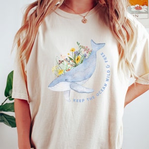 以下が含まれることがあります： クリーム色のTシャツに、青いクジラのイラストが描かれています。クジラはカラフルな花に囲まれており、「Keep the ocean wild & free」という文字が書かれています。