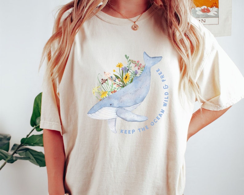 以下が含まれることがあります： クリーム色のTシャツに、青いクジラのイラストが描かれています。クジラはカラフルな花に囲まれており、「Keep the ocean wild & free」という文字が書かれています。