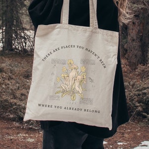 Könnte beinhalten: Eine beige Tragetasche mit schwarzem Text, der "There are places you haven't been where you already belong" lautet. Die Tasche zeigt eine Vintage-Illustration von drei Figuren mit gelben Blumen.