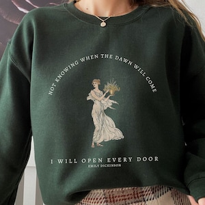 May include: A dark green sweatshirt with a white graphic print featuring a woman holding flowers. The text reads "Not knowing when the dawn will come I will open every door Emily Dickinson".