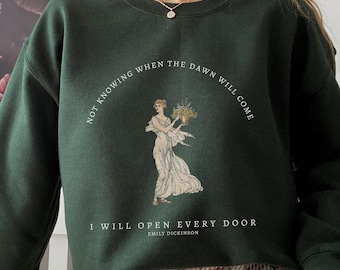 Può includere: Una felpa verde scuro con una stampa grafica bianca che raffigura una donna che tiene dei fiori. Il testo recita "Non sapendo quando arriverà l'alba, aprirò tutte le porte Emily Dickinson".