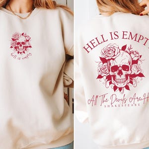 Puede incluir: Una sudadera de color crema con un gráfico rojo de calavera y rosas en la espalda. El texto "El infierno está vacío, todos los demonios están aquí" está impreso debajo del gráfico. El texto "Shakespeare" está impreso debajo de la cita.