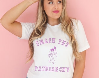 Pode incluir: Camiseta branca com um gráfico roxo de uma mulher jogando tênis e o texto "SMASH THE PATRIARCHY".