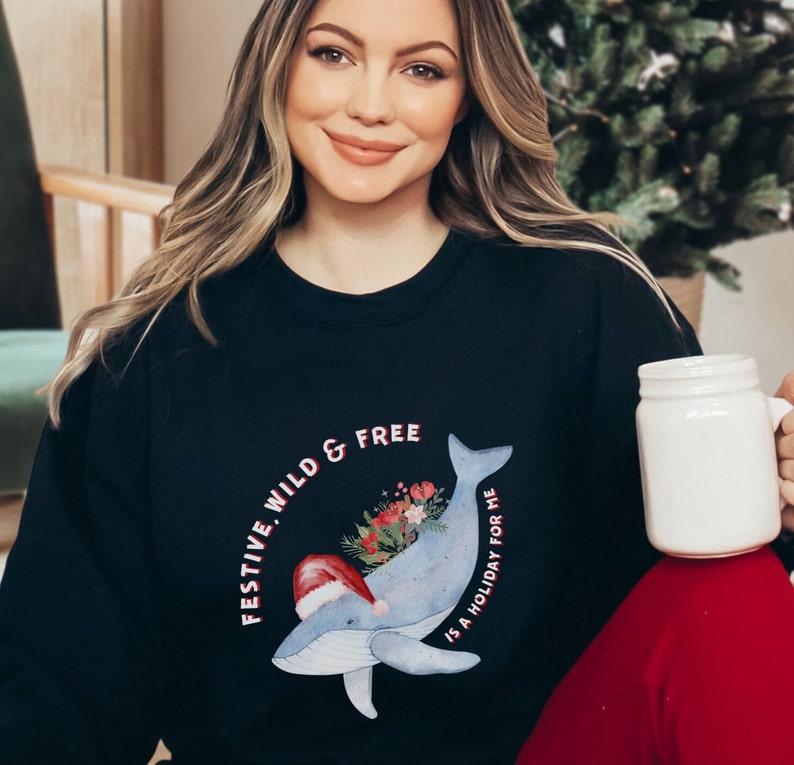 May include: Black sweatshirt featuring a whimsical whale design with a Santa hat and floral accents. The text reads "FESTIVE, WILD & FREE" and "IS A HOLIDAY FOR ME". A festive holiday-themed apparel.