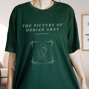 May include: A dark green t-shirt with a white graphic design featuring the title "The Picture of Dorian Gray" by Oscar Wilde. The graphic also includes a line drawing of a face within a decorative frame and the text "To define is to limit."