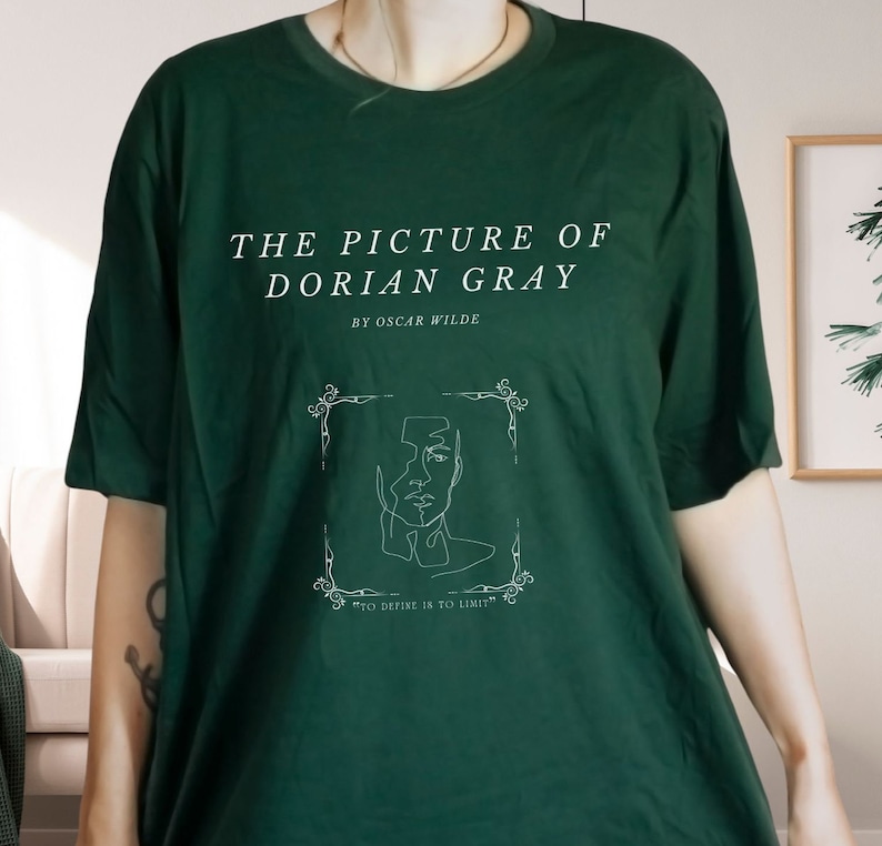 May include: A dark green t-shirt with a white graphic design featuring the title "The Picture of Dorian Gray" by Oscar Wilde. The graphic also includes a line drawing of a face within a decorative frame and the text "To define is to limit."