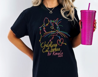 May include: Black t-shirt with a colorful outline of a cat's face and the text "Childless Cat Ladies for Kamala 2024". The design features a heart and is printed in yellow, pink, blue, and green. A pink studded tumbler is held in the hand.