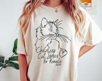 Op de afbeelding: Crèmekleurig T-shirt met een zwarte lijntekening van een kat. De tekst "Childless Cat Ladies for Kamala 2024" staat eronder, met een hartsymbool en het jaartal.