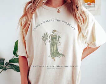 May include: A cream-colored t-shirt with a vintage illustration of a woman in a forest. The text "I took a walk in the woods and came out taller than the trees" is printed on the shirt, along with the author's name, "Henry David Thoreau".