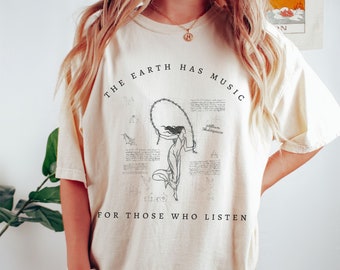 May include: A cream colored t-shirt with a black graphic print that reads "The Earth Has Music For Those Who Listen". The graphic features a woman holding a rope and a quote from William Shakespeare.