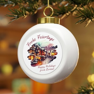 May include: White Christmas bauble featuring a watercolour illustration of a snowy village scene. The bauble has the text "Frohe Feiertage" and "Happy Holidays from Zermat" printed on it.