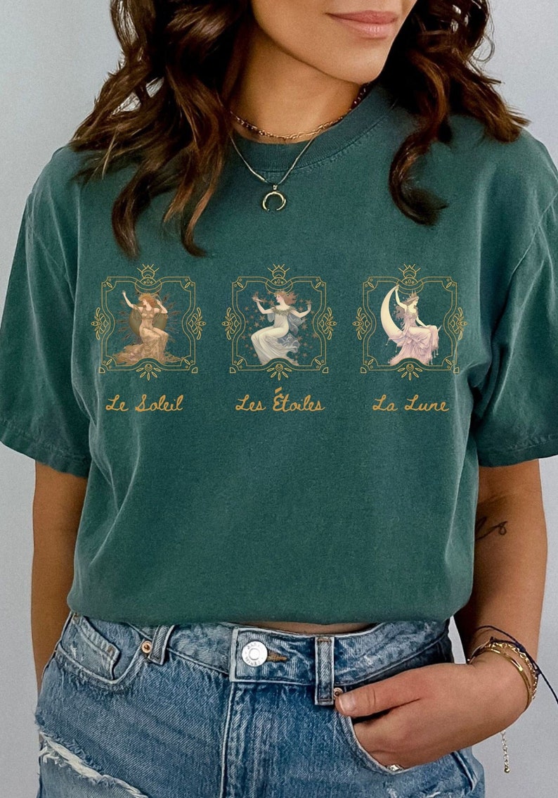 May include: A teal green t-shirt with a graphic print of three women in gold frames. The text below the frames reads "Le Soleil", "Les Etoiles", and "La Lune".