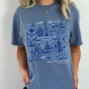 K&ouml;nnte beinhalten: Ein blaues T-Shirt mit einem wei&szlig;en und blauen Delft, Zuid-Holland, Niederlande Design. Das Design zeigt eine Vielzahl traditioneller niederl&auml;ndischer Bilder, darunter H&auml;user, Windm&uuml;hlen und Blumen.