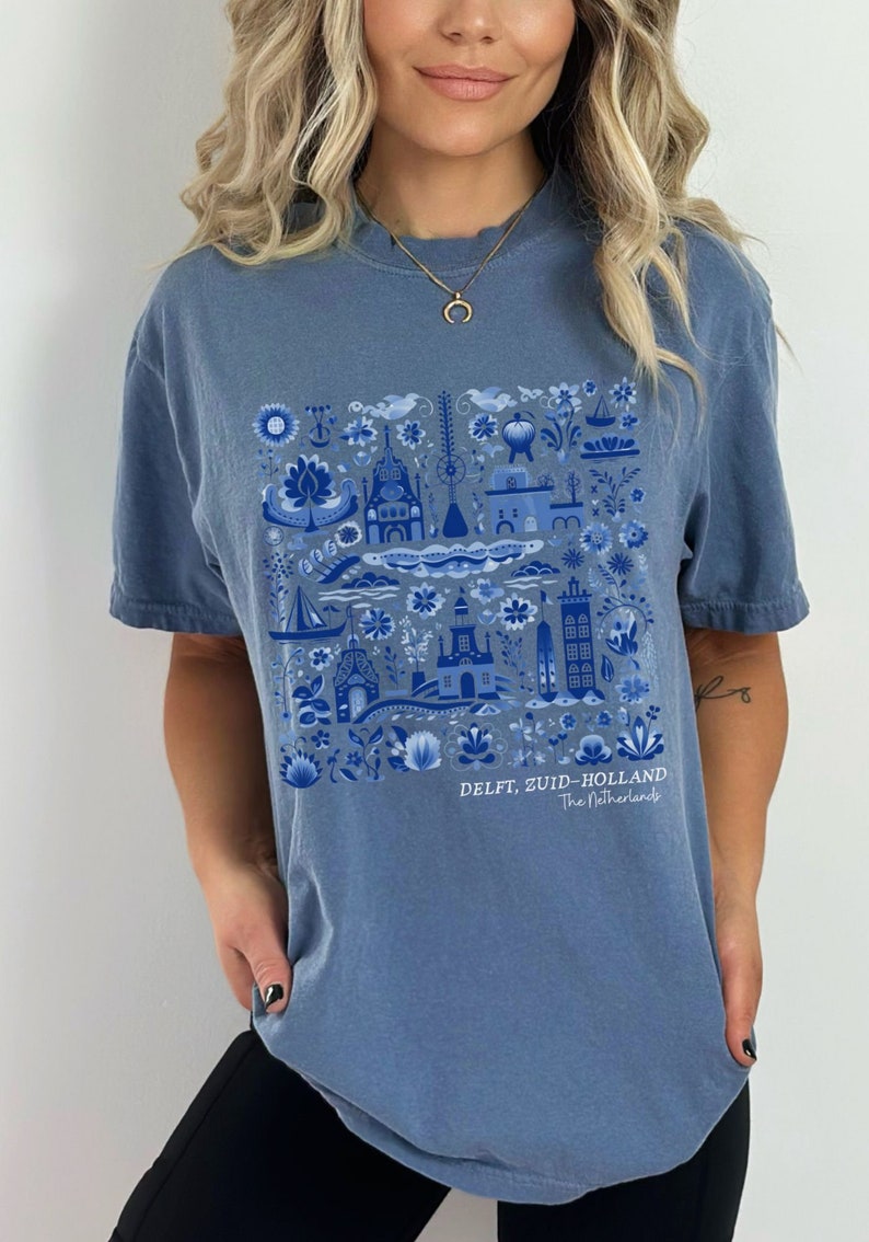 K&ouml;nnte beinhalten: Ein blaues T-Shirt mit einem wei&szlig;en und blauen Delft, Zuid-Holland, Niederlande Design. Das Design zeigt eine Vielzahl traditioneller niederl&auml;ndischer Bilder, darunter H&auml;user, Windm&uuml;hlen und Blumen.