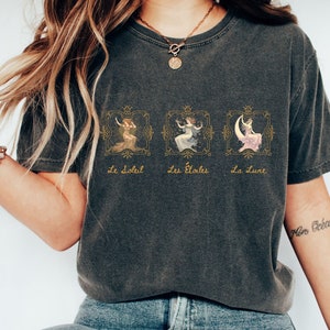 May include: A dark gray t-shirt with a graphic print of three women in ornate frames. The text "Le Soleil", "Les &Eacute;toiles", and "La Lune" is printed below the frames. The shirt has a cropped fit and is worn by a person with long blonde hair.