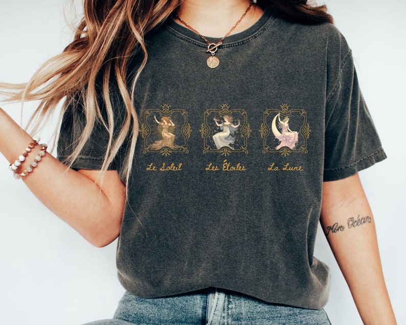 May include: A dark gray t-shirt with a graphic print of three women in ornate frames. The text "Le Soleil", "Les &Eacute;toiles", and "La Lune" is printed below the frames. The shirt has a cropped fit and is worn by a person with long blonde hair.