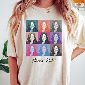 以下が含まれることがあります： カラフルなポップアートのデザインが施された白いTシャツ。女性の顔が9回繰り返されています。デザインの下には「Harris 2024」という文字が印刷されています。