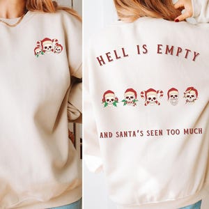 Puede incluir: Una sudadera de color crema con el texto "Hell is empty and Santa's seen too much" impreso en la espalda. La parte delantera de la sudadera presenta un pequeño diseño de cinco calaveras con temática navideña.