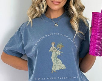 May include: A blue t-shirt with a graphic of a woman holding flowers and the text "Not knowing when the dawn will come I will open every door".