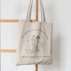 May include: A beige tote bag with a quote by Emily Dickinson printed on it. The quote reads "Not knowing when the dawn will come I will open every door". The bag features a vintage illustration of a woman holding a bouquet of flowers.