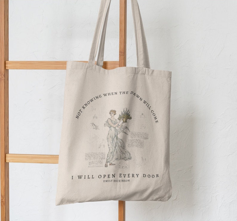 May include: A beige tote bag with a quote by Emily Dickinson printed on it. The quote reads "Not knowing when the dawn will come I will open every door". The bag features a vintage illustration of a woman holding a bouquet of flowers.