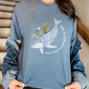 以下が含まれることがあります： 花柄のクジラが描かれた青いTシャツ。Tシャツには「Keep the ocean wild & free」の文字がプリントされています。