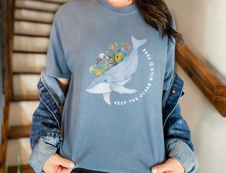以下が含まれることがあります： 花柄のクジラが描かれた青いTシャツ。Tシャツには「Keep the ocean wild & free」の文字がプリントされています。