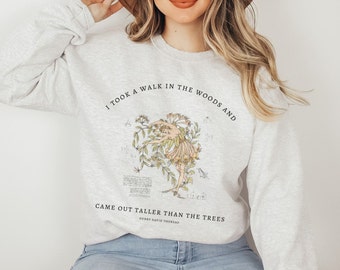 May include: A gray sweatshirt with a graphic print of a woman dancing in a forest. The text on the sweatshirt reads "I took a walk in the woods and came out taller than the trees" and is attributed to Henry David Thoreau.