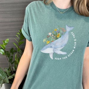 以下が含まれることがあります： 青いクジラと花のイラストが描かれたティールグリーンのTシャツ。クジラは花に囲まれ、「Keep the ocean wild & free」という文字が書かれています。