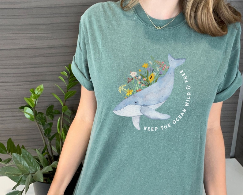 以下が含まれることがあります： 青いクジラと花のイラストが描かれたティールグリーンのTシャツ。クジラは花に囲まれ、「Keep the ocean wild & free」という文字が書かれています。