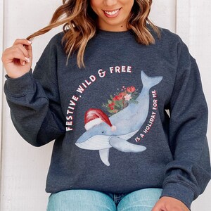 May include: Dark gray sweatshirt with a whimsical whale design. The whale is light blue and wears a Santa hat. The whale has a floral arrangement on its back. Text reads "FESTIVE, WILD & FREE IS A HOLIDAY FOR ME."
