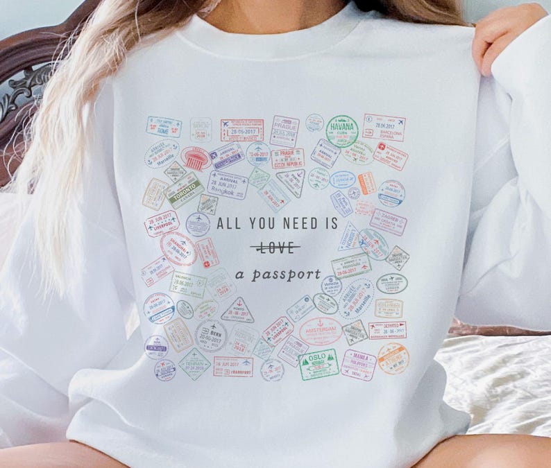 Pode incluir: Camisola branca com um gr&aacute;fico de um cora&ccedil;&atilde;o feito de carimbos de passaporte. O texto "All you need is love - a passport" est&aacute; impresso abaixo do cora&ccedil;&atilde;o.