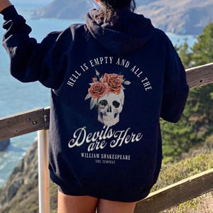 Puede incluir: Sudadera con capucha negra con una calavera que lleva una corona de flores. El texto en la sudadera dice "El infierno está vacío y todos los demonios están aquí. William Shakespeare La Tempestad."